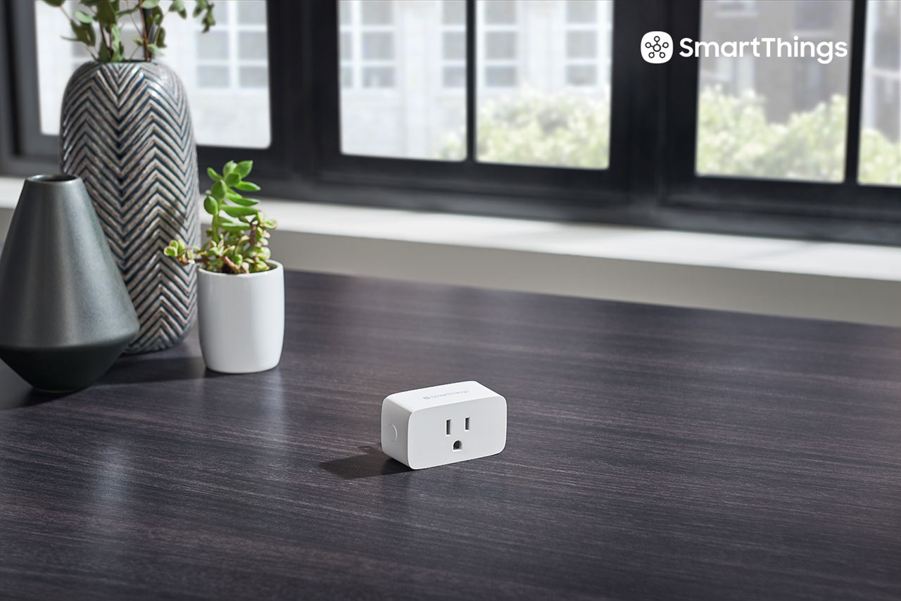  A smart plug placed on a desk. Behind it are some decorative potted plants  